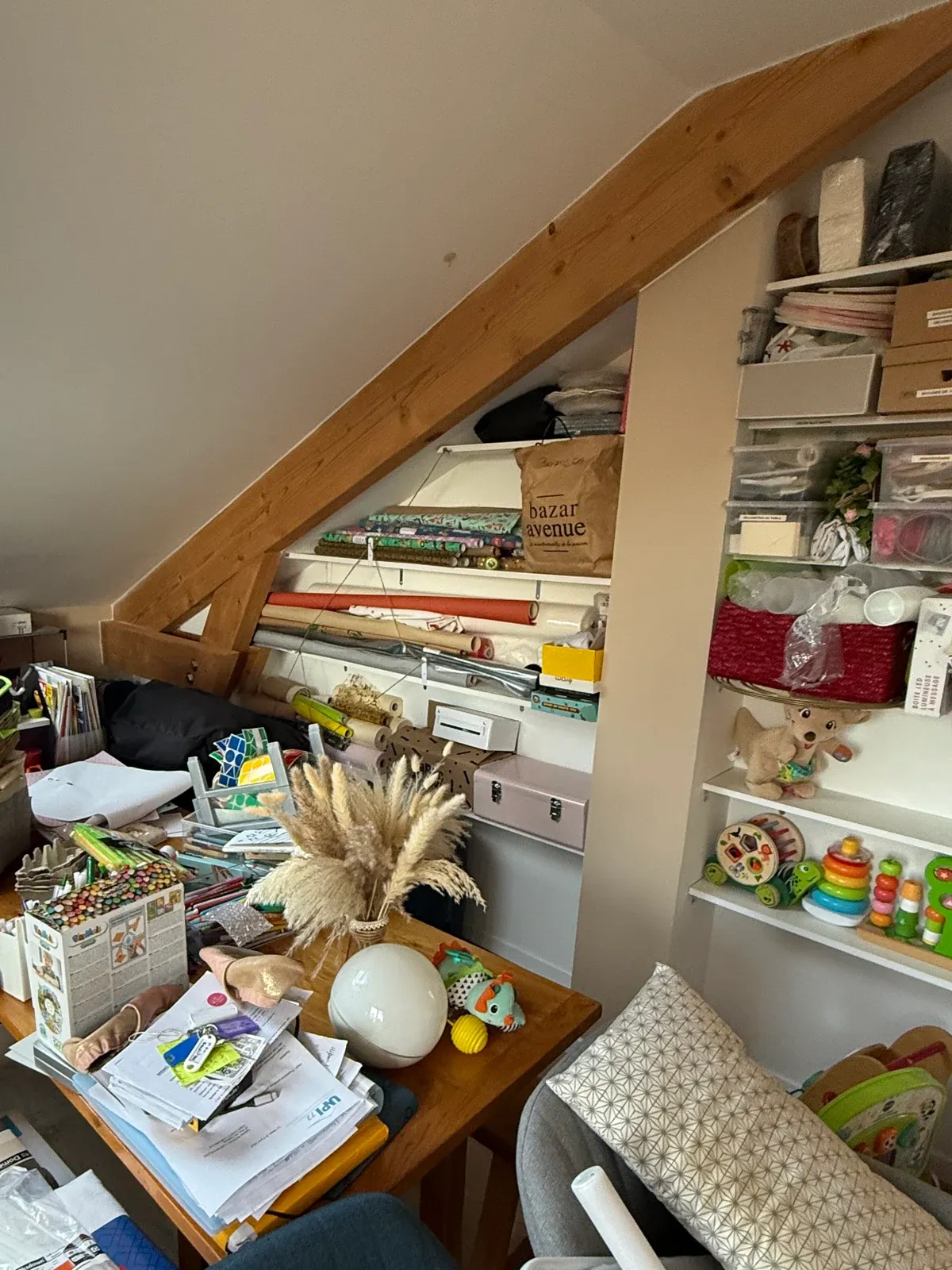 Table de travail du bureau sous les combles couverte de papiers et d'objets, devant des étagères chargées