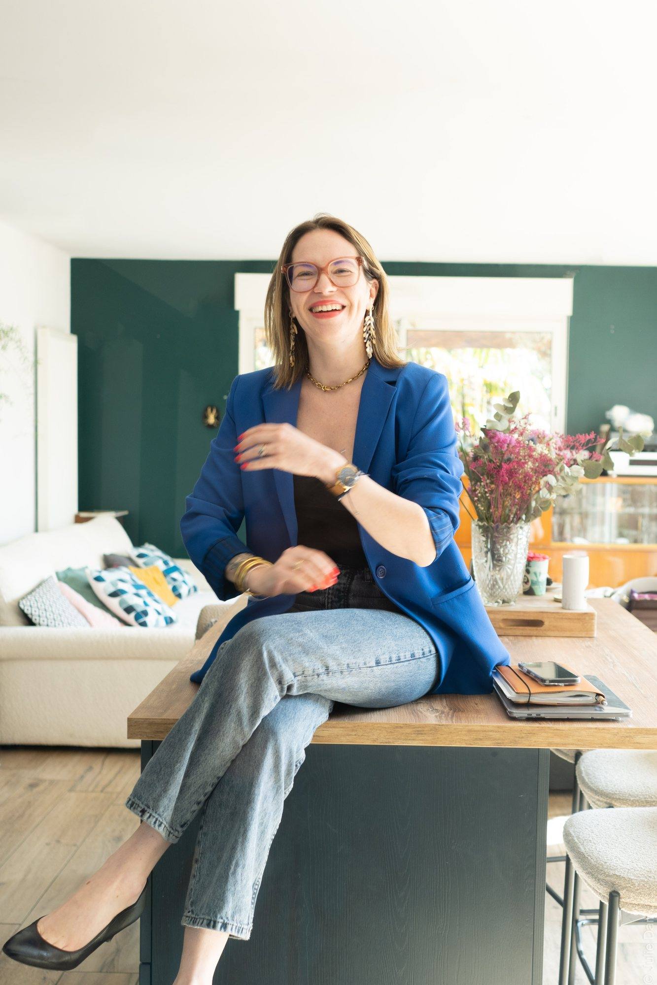 Virginie Pinsault en blazer bleu, assise sur une table en bois dans un salon au mur vert